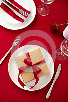 Valentine`s day dining table concept. Plate with gift box, utensil, bottle of wine on red background. Flat lay, top view