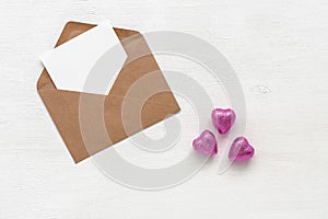 Valentine`s Day. Greeting blank card mockup in an envelope. Chocolate candies in a heart shape in a purple wrapper. White wooden