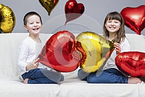 Valentine`s Day, a holiday for all lovers. Cute Caucasian children, boy and girl are sitting on the sofa with red and gold heart-