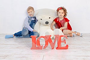 Valentine`s day kids. Boy and girl sit with a big teddy bear, the inscription love in focus