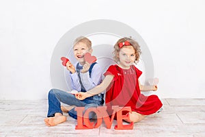 Valentine`s day kids. A boy and a girl sit with a large inscription Love and hold hearts in their hands