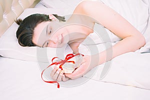 Valentine`s day morning. Beautiful brunette girl lying on the bed holding a white gift box with a red ribbon at home