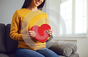 Valentine`s day. Red heart in the hands of a girl in casual clothes sitting on the couch