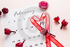 Valentine`s Day. A red heart-shaped ribbon highlights the date February 14 on a calendar sheet and dried flowers on a pink