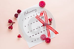 Valentine`s Day. A red heart-shaped ribbon highlights the date February 14 on a calendar sheet and dried flowers on a pink
