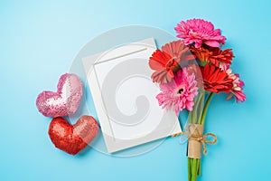 Valentine`s day white picture frame mock up with gerbera daisy flower bouquet and heart shapes  on blue background. Top view, fla