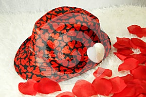 Valentines black hat with many red hearts, on white shag carpet, rose petals, small white pillow hat