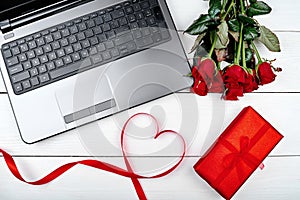 Valentines Day background with bouquet of red roses, ribbon shaped as heart, gift box and open laptop computer, copy space. Love