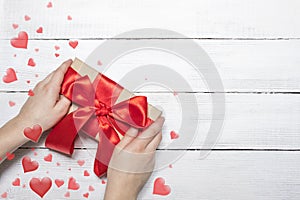 Valentines day background. Hands hold a box with a gift and a red ribbon on a white wooden background, hearts fly rippled. The