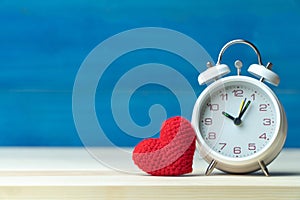 Valentines day concept. hand make yarn red heart in front of white alarm clock on wooden table and blue background