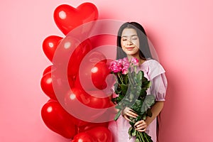 Valentines day concept. Romantic teen asian girl dreaming of love or date, close eyes and smile, holding flowers from