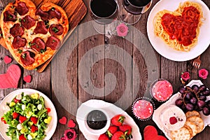 Valentines Day dinner frame on a dark wood background with heart shaped pizza, pasta, wine, cheese plate and desserts