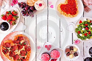 Valentines Day dinner frame on a white wood background with heart shaped pizza, pasta, wine, cheese plate and desserts