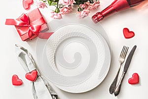 Valentines day dinner with table place setting with red gift, a bottle of champagne, pink roses, hearts with silverware on white. photo