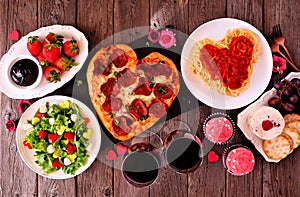 Valentines Day dinner table scene on dark wood with heart shaped pizza, pasta, wine, cheese plate and desserts