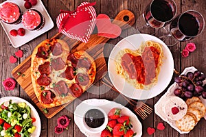 Valentines Day dinner table scene on dark wood with heart shaped pizza, pasta, wine, cheese plate and desserts