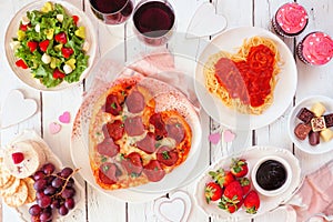 Valentines Day dinner table scene on white wood with heart shaped pizza, pasta, wine, cheese plate and desserts