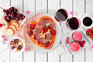 Valentines Day dinner table scene on white wood with heart shaped pizza, wine, cheese plate and desserts