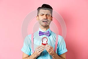 Valentines day. Heartbroken guy crying and holding engagement ring, sobbing from break-up, standing over pink background