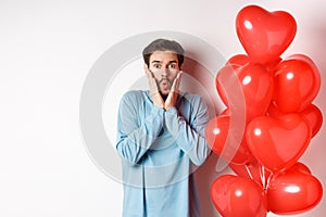 Valentines day. Image of young man standing near hearts balloons with shocked face, staring startled at camera, white