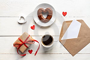 Valentines day mockup. Coffee, heart shaped chocolate cookie, gift box and paper card with craft envelope on white wooden table