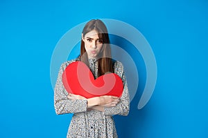 Valentines day. Sad and gloomy caucasian girl pucker lips and looking disappointed, holding big red heart cutout