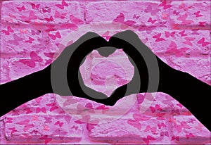 Valentines day, silhouette of two hands making a heart shape together on a pink brick wall with butterfly`s