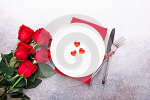 Valentines day table place setting with bouquet of roses, red hearts and silverware on stone background. Top view. Valentine`s