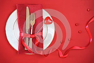 Valentines day table setting with white plate fork and knife shape heart on red tablecloth with ribbon bow background. Romantic