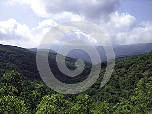 Valley of hills covered with low green forest. In the distance, the silhouettes of high mountains turn blue. Clear blue sky with