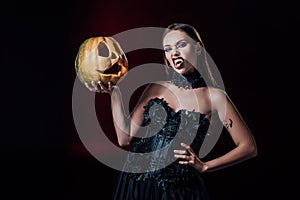 Vampire girl with fangs in black gothic dress holding Halloween pumpkin on black background