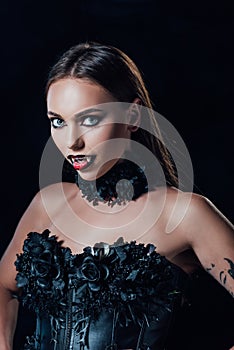 Vampire girl with fangs in black gothic dress looking at camera isolated on black