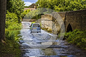 A van crosses the ford of the bridge and ford over the River Ise in the town of Geddington, UK