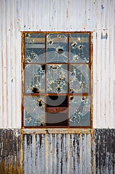 Vandalized window in old tin industrial building with holes from thrown rocks and or bullets in reinforced panes in rusty frame