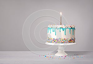 Vanilla Birthday Cake with a lit gold candle, teal blue drip and colorful sprinkles on a light grey white background