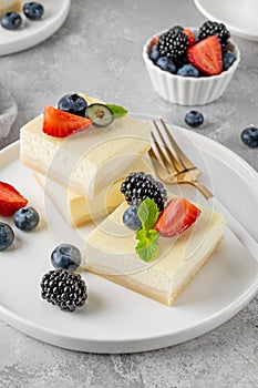 Vanilla cheesecake cut into squares with fresh berries on a white plate on a gray stone background