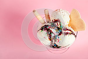 Vanilla flavor ice cream in glass bowl with chocolate sauce, strewed sprinkles and waffles on pink background