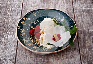 Vanilla ice cream with mint and pretty flower. A bowl of icecream and pink raspberries on a wooden background. Cold summer snacks.