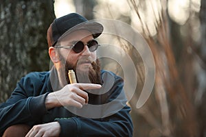 Vape. A brutal young man with a huge beard in sunglasses and a cap and with electronic cigarette in the woods at sunset.