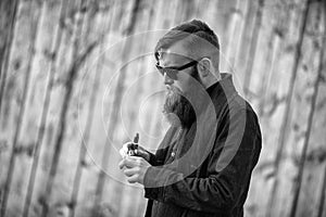 Vape man. Outdoor portrait of a young brutal white guy with large beard vaping electronic cigarette opposite the old wooden fence.