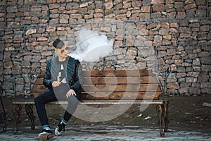 Vape. A young handsome guy sits on the bench and blows steam from an electronic cigarette.