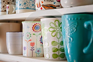 A variety of ceramic mugs and cups in spring floral bright style stand in a row on a white home shelf.