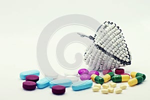 A variety of different coloured pills strewn around a white beaded wire heart. Set against a near-white background and surface.