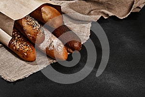 Variety of fresh baked bread in a paper bag, laying on a burlap against black background, copy space. Close-up