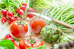 Variety of Fresh Organic Vegetables Artichokes Green Beans Tomatoes Red Radish Broccoli on Wood Garden Kitchen Table in Sunlight