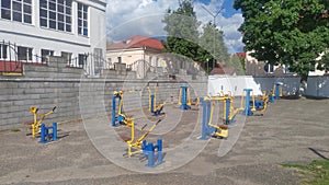 A variety of metal exercise equipment for general use is set up on an asphalted area along a concrete fence. Nearby, trees grow an