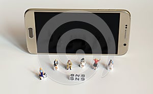 A variety of miniature people sitting with  `SNS` alphabetical white cube in front of Smart phone.