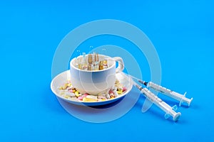 A variety of pills in a coffee cup, syringes next to a blue background close-up