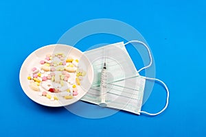 A variety of pills on a plate, medical masks and syringes next to each other on a blue background close-up