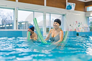 Variety of swimming equipment. Green foam noodles used by active happy caucasian senior couple. Swimming pool interior.
